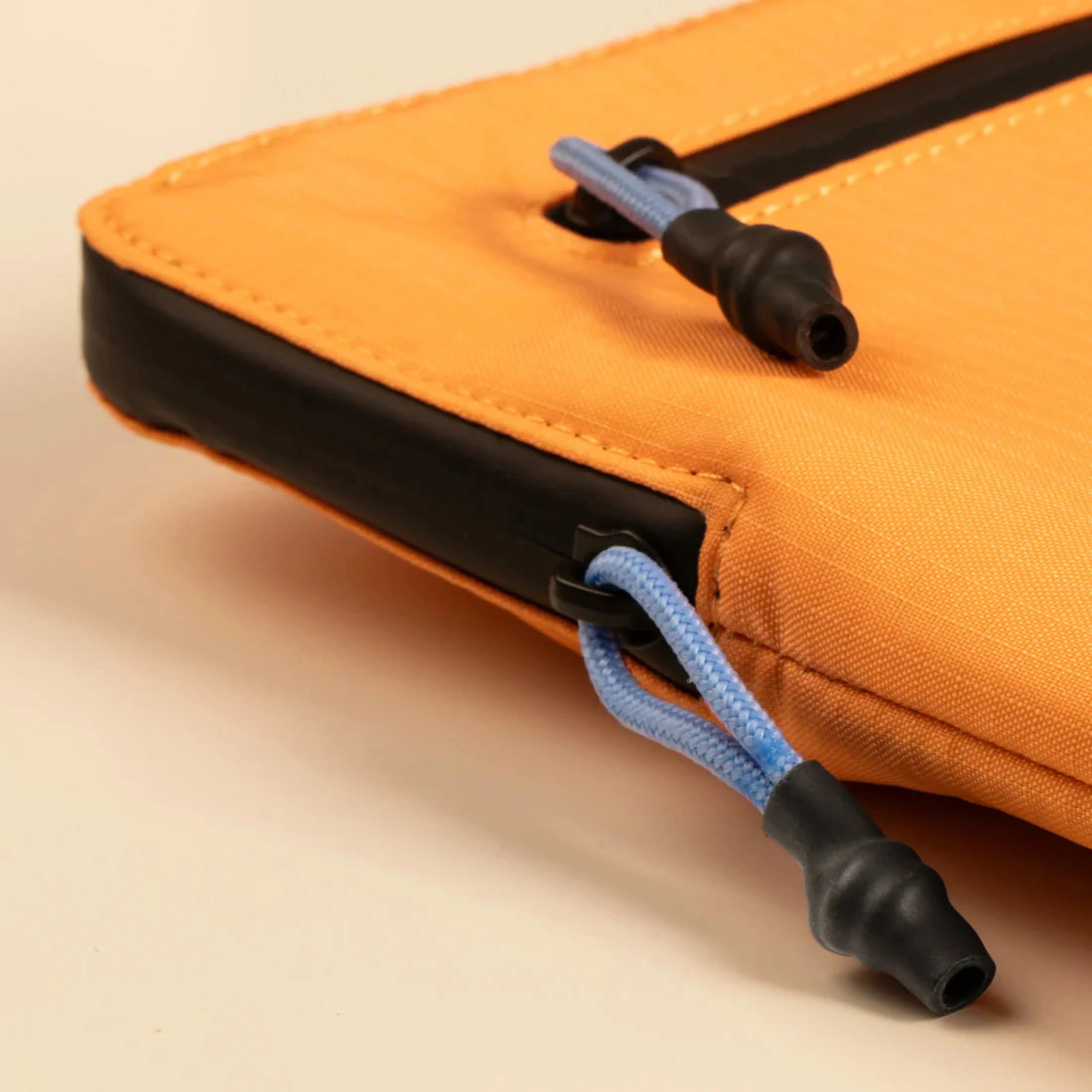 Close-up of an orange technical bag showing blue bungee zipper pulls and reinforced edge detailing