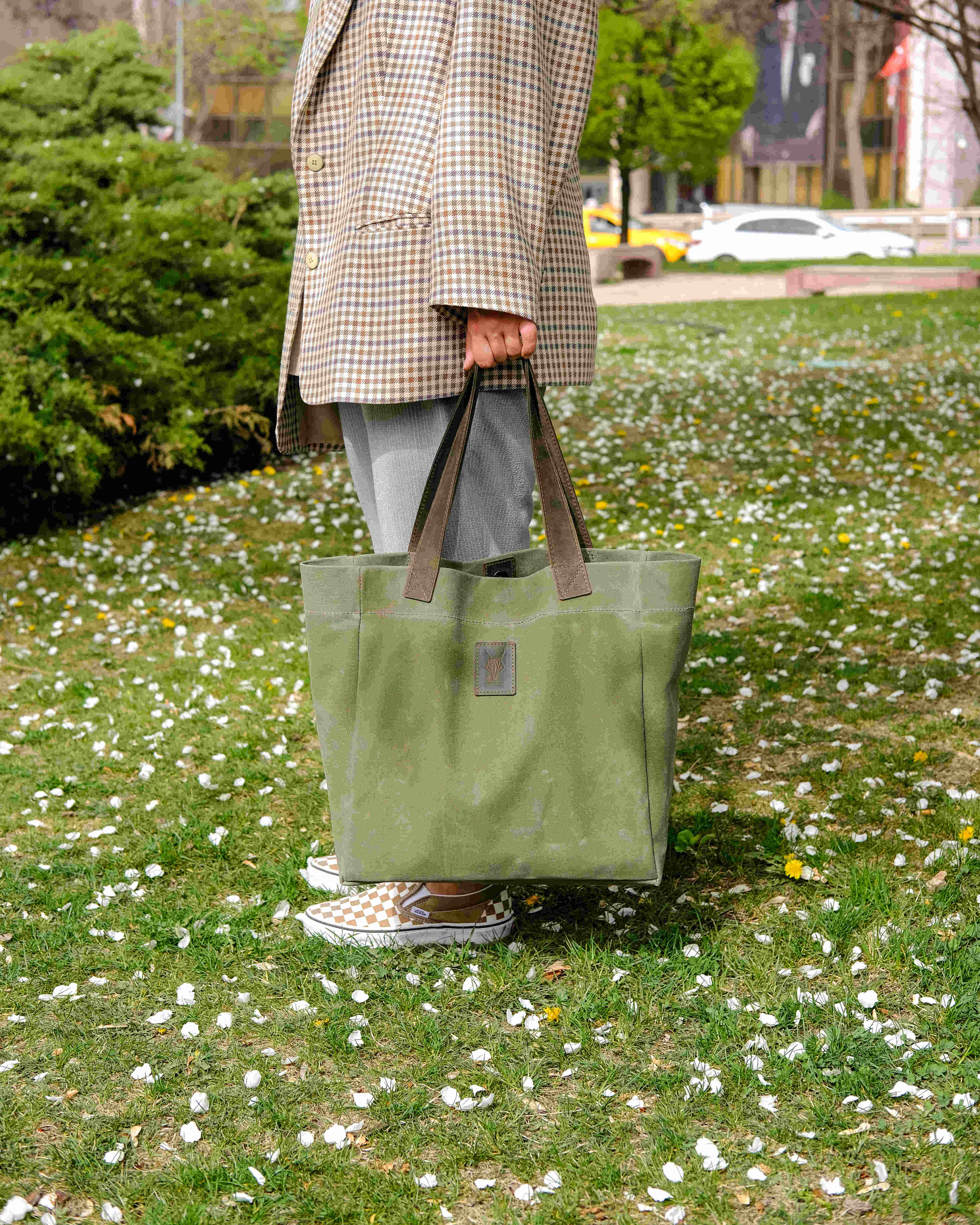 Olive canvas tote bag with sturdy handles held by a person outdoors on grass with scattered white petals